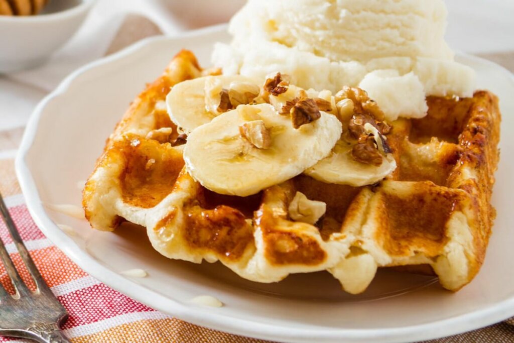 Waffle de banana com nozes em um prato branco com uma bola de sorvete, duas rodelas de banana e nozes em cima