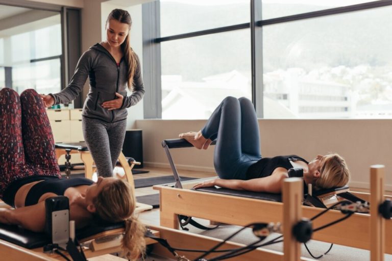Duas mulheres com roupas de academia deitadas em aparelho realizando exercício de pilates acompanhadas de professora de pilates usando roupas de academia na cor cinza