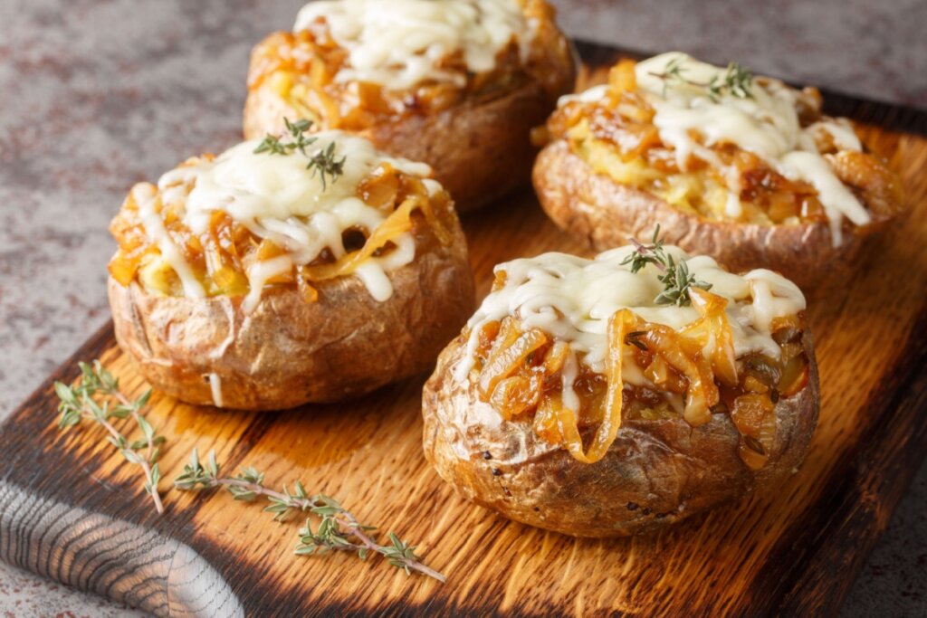 Quatro batatas recheadas com cebola caramelizada e queijo em cima de uma tábua de madeira