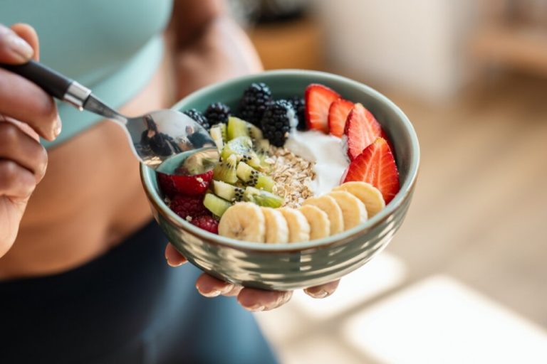 Mãos segurando uma colher e um recipiente verde com banana, morango, kiwi, framboesa, amora, flocos de aveia e iogurte