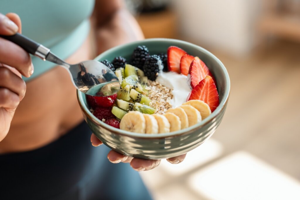 Mãos segurando uma colher e um recipiente verde com banana, morango, kiwi, framboesa, amora, flocos de aveia e iogurte