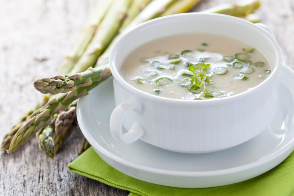 recipiente e prato branco com sopa clara com aspargo em cima de pano verde. aspargos ao lado