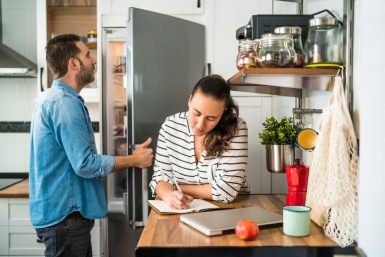 Casal na cozinha montando lista de compras, o homem está usando uma camisa jeans e calça preta e conferindo os alimentos na geladeira e a mulher está fazendo a lista de compras, ela está com o cabelo preso e usando uma camisa de manga longa listrada