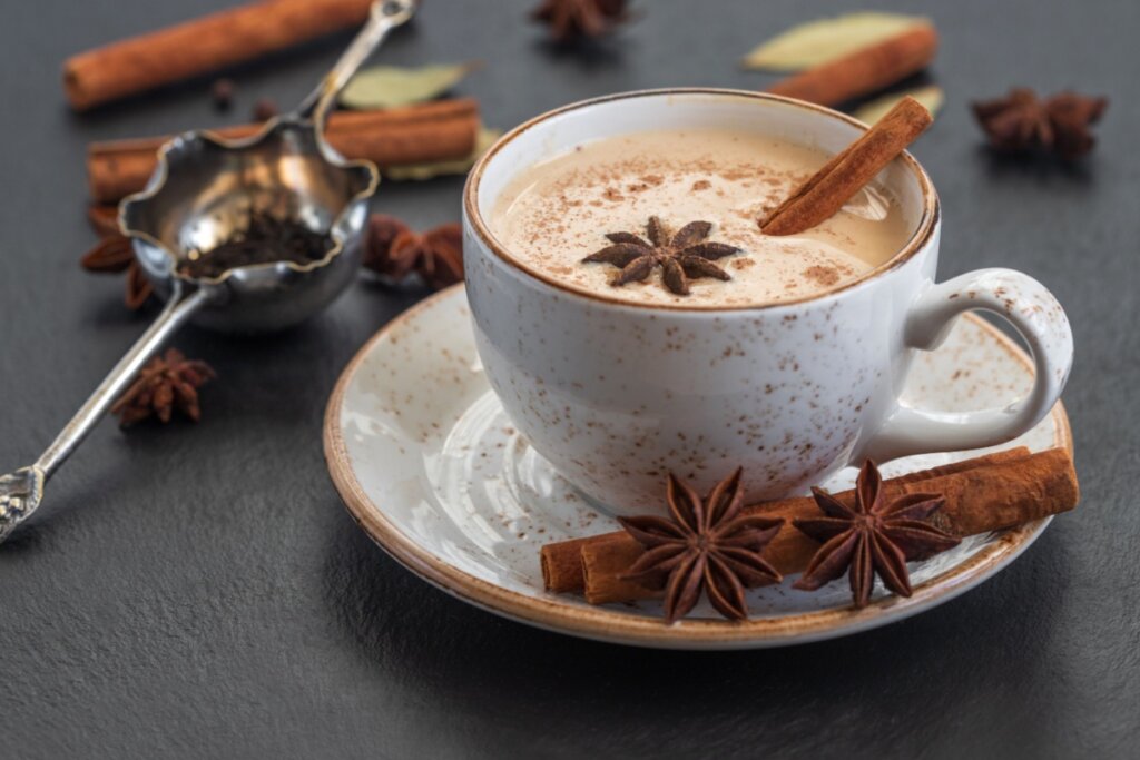 Chai latte servido em xícara estampada de cerâmica em cima de mesa de madeira preta com castanha e cravos-da-índia.