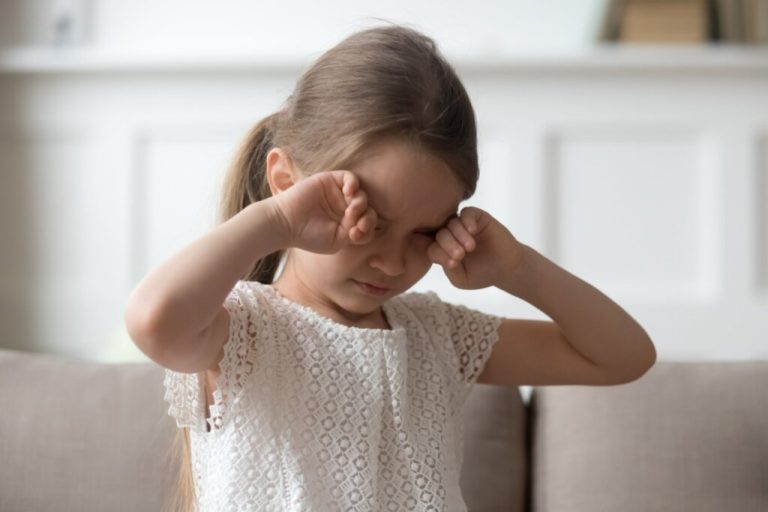 Menina com as duas mãos nos olhos, como se estivesse com sono, vestindo uma blusa branca e sentada em um sofá