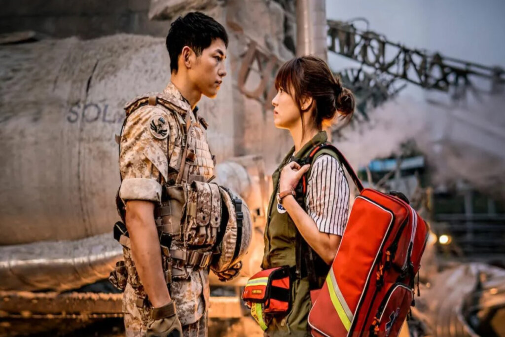 Imagem de um homem com roupa de soldado de frente para uma mulher usando roupas e equipamentos de resgate ao fundo uma construção destruída