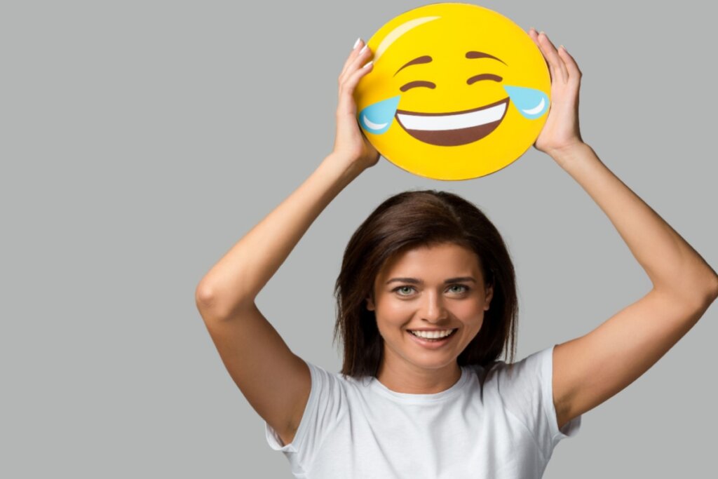 Mulher sorridente segurando acima da cabeça um grande emoji amarelo de rosto rindo com lágrimas, sobre fundo cinza