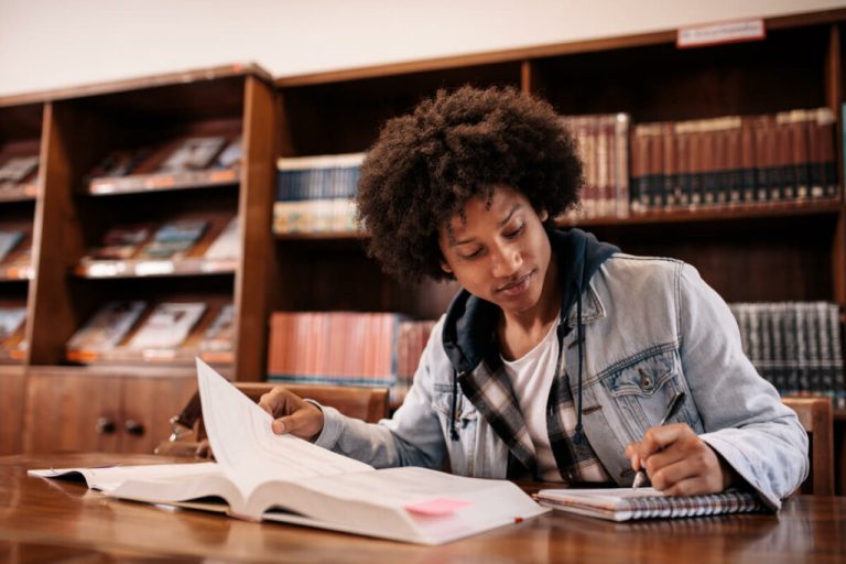Menino sentado estudando com cadernos e livros abertos em cima da mesa e uma biblioteca ao fundo