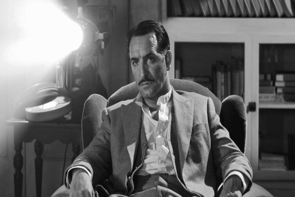 Imagem em preto e branco de um homem com cabelo curto e bigode sentado em poltrona com roupa social assistindo a projeção