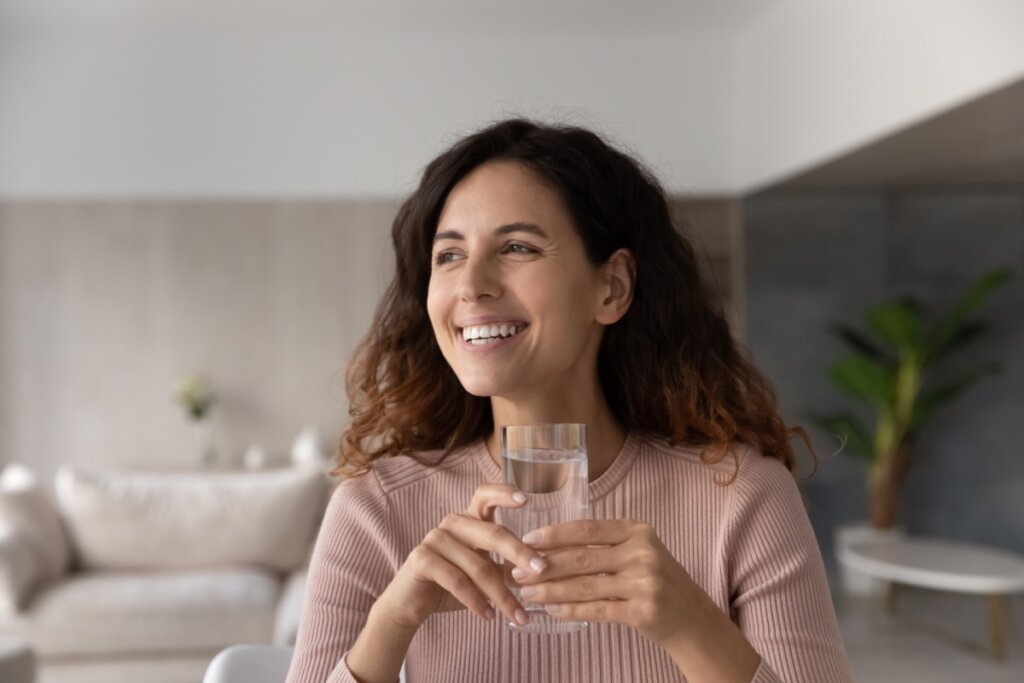 Mulher sorrindo, sentada e segurando um copo de vidro com água