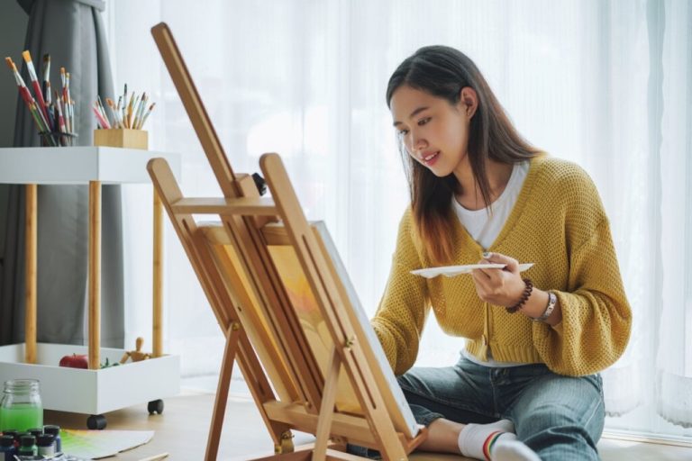 Jovem com o cabelo liso, escuro e solto. Usando uma camiseta branca e um suéter amarelo, calça jeans e uma meia branca com detalhes coloridos, sentada no chão pintando tela em cavalete