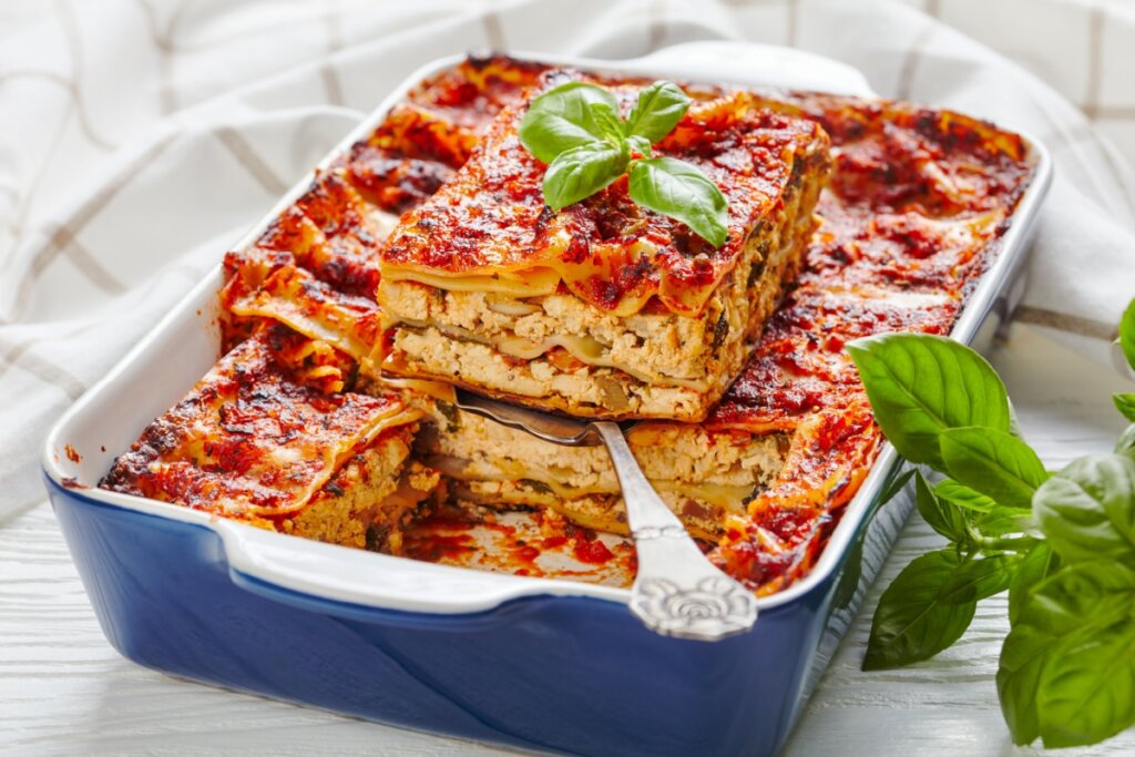 Lasanha de cogumelo com tofu servida em tigela de cerâmica azul, em cima de mesa de madeira branca