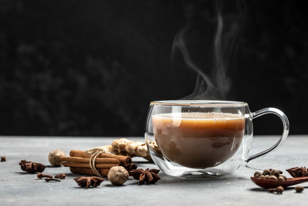 Masala chai servido em xícara de vidro em cima de mesa de madeira com os ingredientes ao redor da xícara como canela, cravos da índia