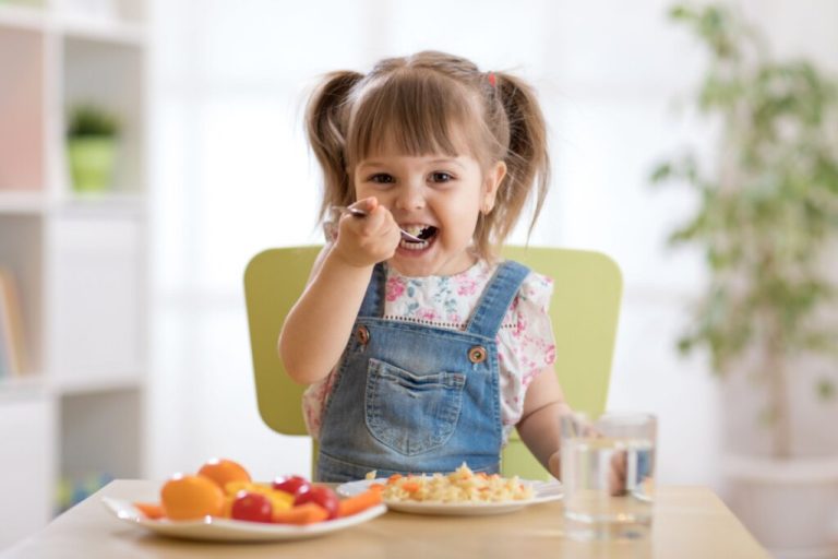 Menina sentada em mesa com dois pratos com alimentos e copo de água. ela está com colher na boca
