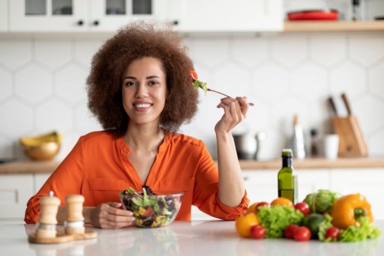 Mulher com cabelo cacheado usando camisa laranja, sentada fazendo refeição saudável, segurando uma tigela de salada em uma mão e na outra um garfo 