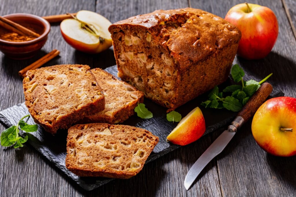 Pão doce de maçã fatiado, com pedaços visíveis da fruta e toque de canela, servido sobre tábua rústica com maçãs frescas ao redor