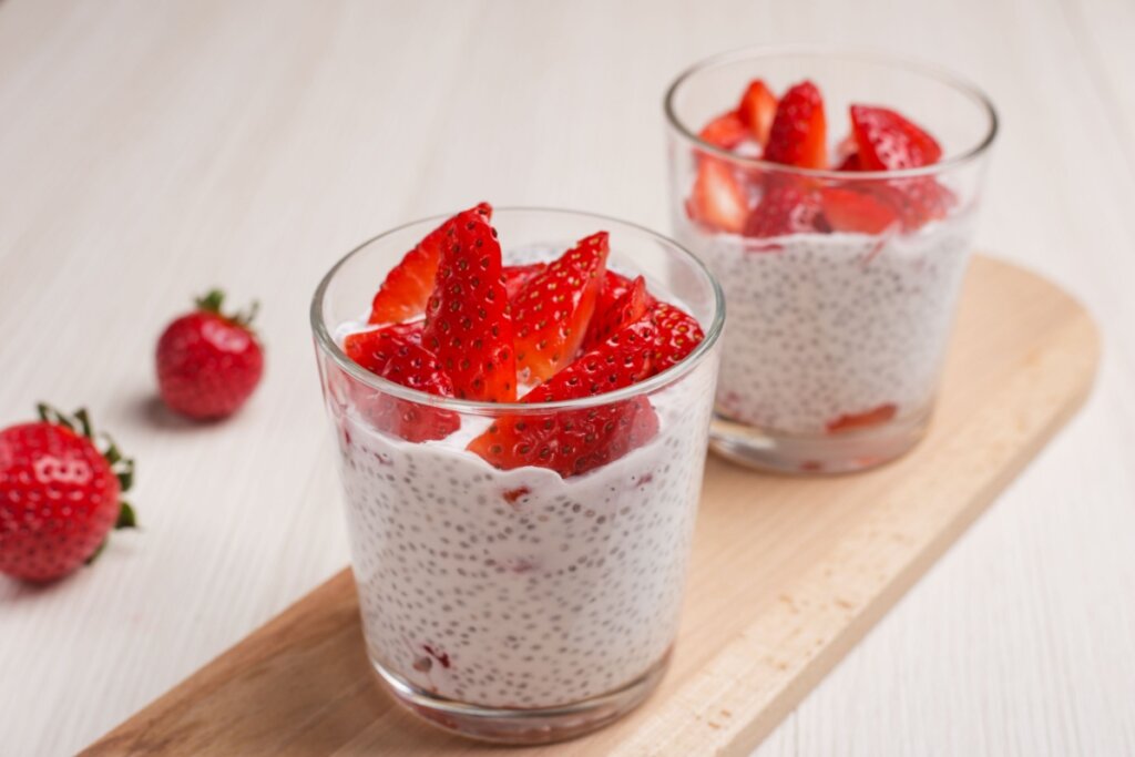Dois copos com pudim de chia com linhaça e com morangos cortados em cima do pudim. Em cima de uma bandeja na bancada e morangos ao lado