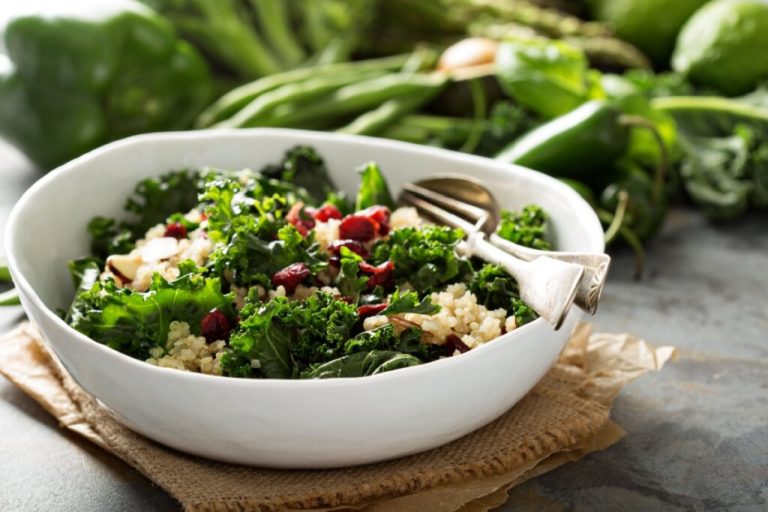 prato branco em cima de pano marrom com salada de couve com quinoa, cranberries e amêndoas. alimentos verdes desfocados ao fundo