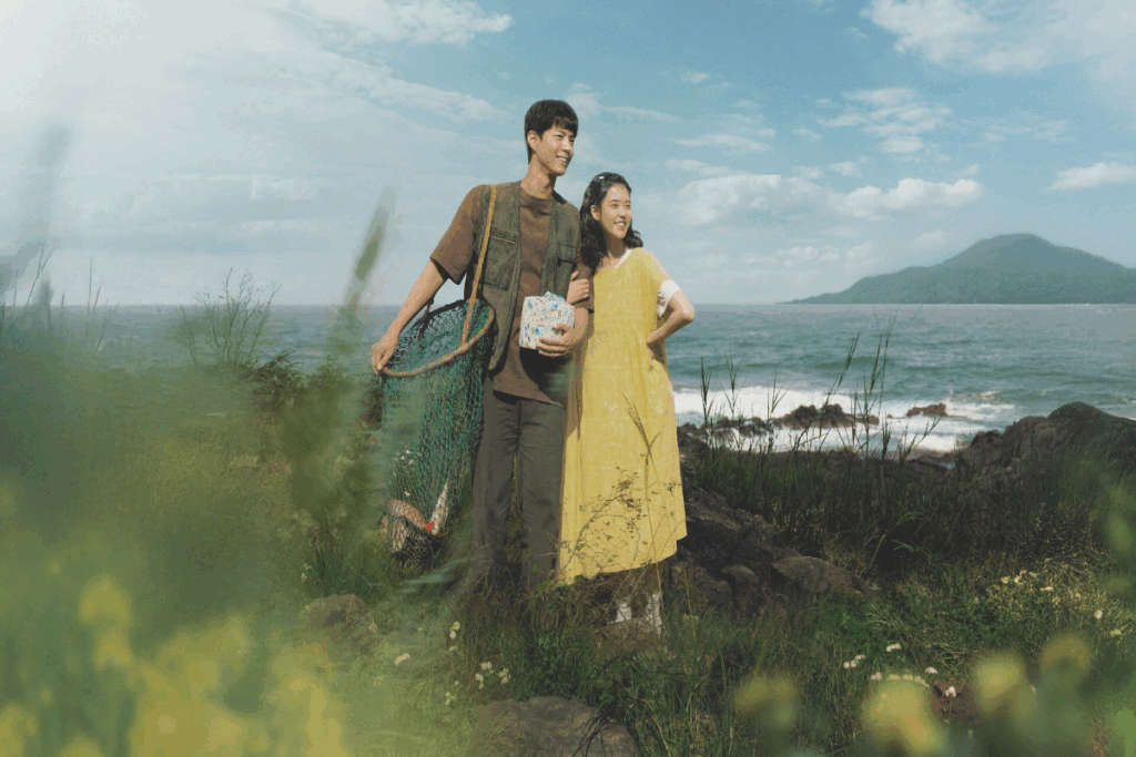 Casal em campo florido abraçado, o homem está usando roupas simples de pescador com uma rede de peixes no ombro e a mulher está usando um vestido amarelo com a mão na cintura