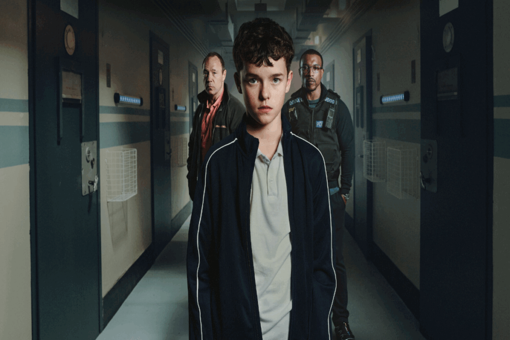 Adolescente com o cabelo preto e curto, usando camiseta cinza e casaco azul-escuro com uma listra branca e atrás estão dois homens, um com uma camisa vermelha e casaco escuro e o outro com uma roupa de policial