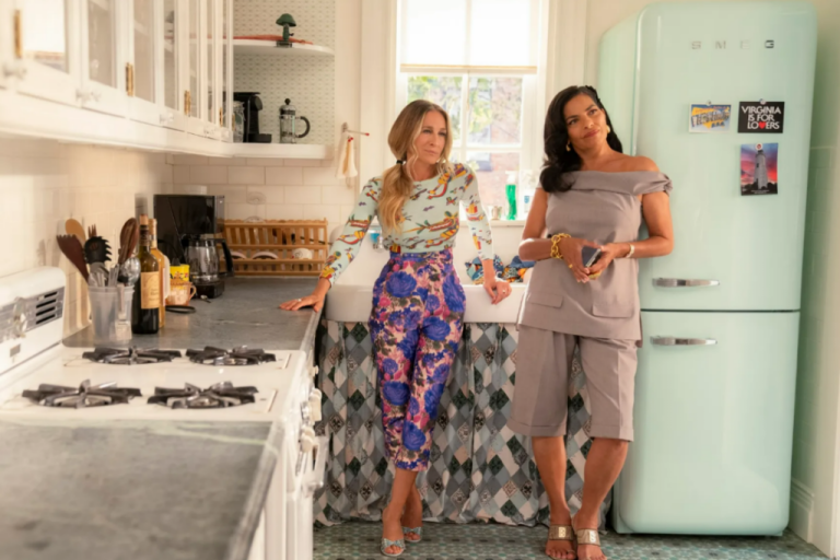 Duas mulheres em uma cozinha decorada com moveis brancos e uma geladeira verde-água, usando roupas coloridas