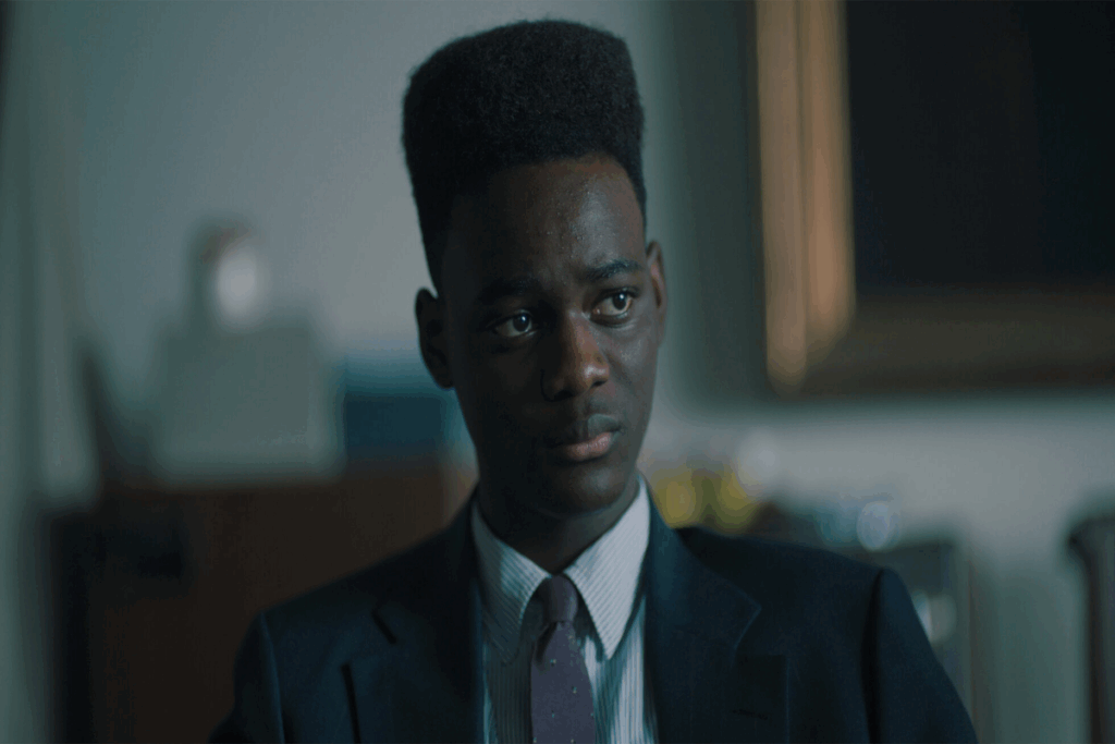 Jovem negro com o topete usando camisa social branca, gravata cinza e terno preto com uma expressão aflita
