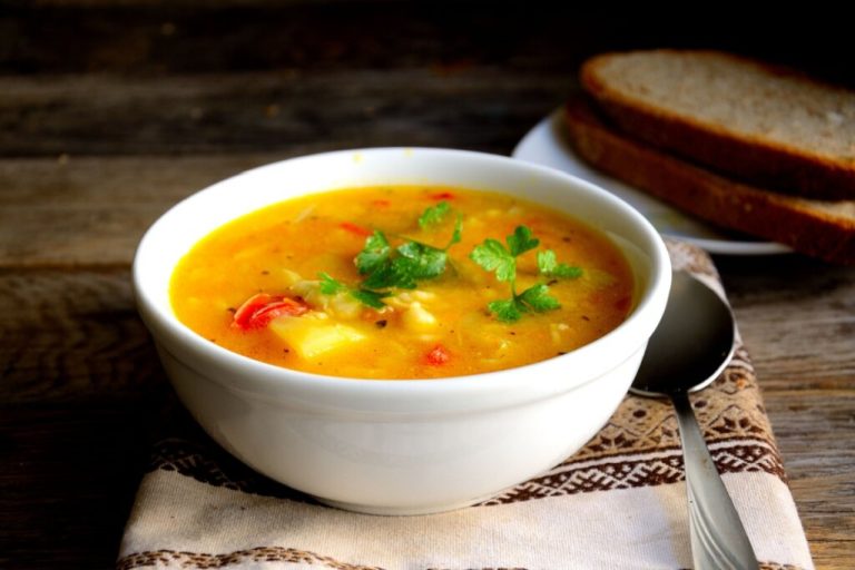 recipiente branco em cima de pano com sopa com caldo laranja e legumes, como cenoura, batata e pimentão vermelho, decorada com salsinha
