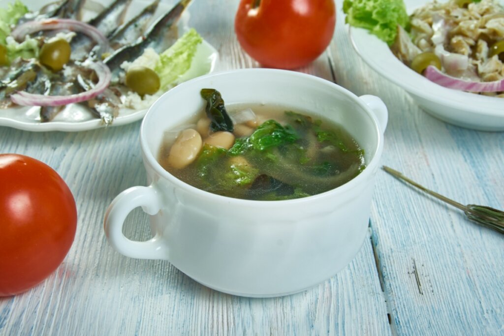 Mini panela branca com sopa de escarola com feijão-branco dentro em cima de uma mesa de madeira branca