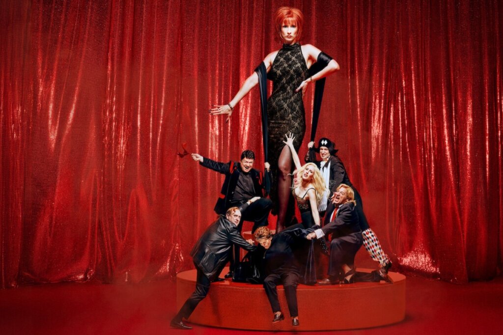 Grupo de personagens cerca uma figura central, Yurena, em um palco vermelho, com cortinas ao fundo