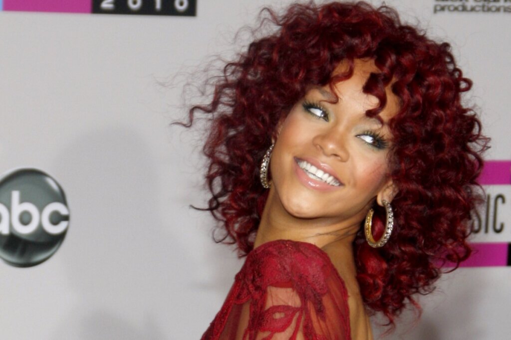 Rihanna usando cabelo cacheado em tom cereja glossy, sorrindo e posando para foto com um vestido vermelho rendado