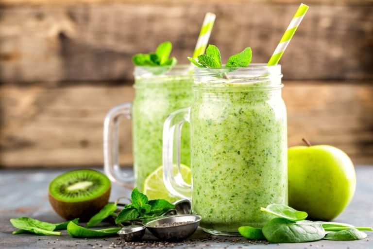 Vitamina de kiwi, espinafre e maça-verde servido em copos de vidro em cima de mesa de madeira, com pedaços de kiwi, folhas de espinafre e uma maça-verde ao redor