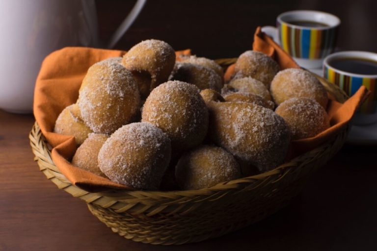 Cesta de vime repleta de bolinhos de chuva dourados e açucarados, acompanhada de xícaras de café