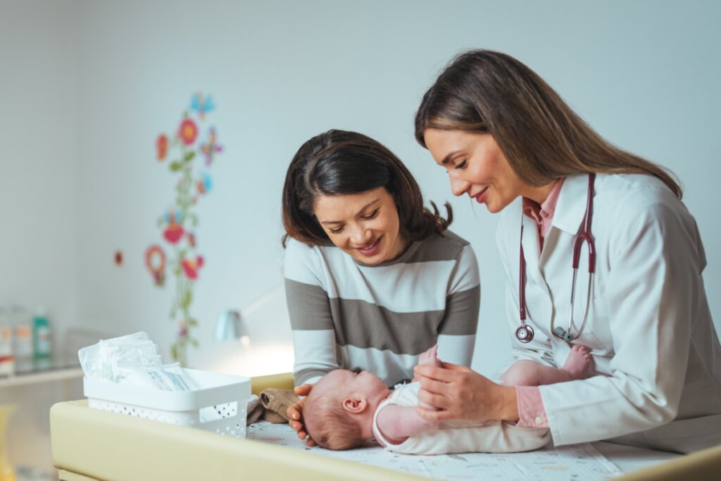 Bebê em consulta com pediatra e com a mãe ao lado. Uma das mulheres tem o cabelo curto e liso, e está usando uma camisa de manga longa listrada e a outra tem o cabelo longo e liso, e está usando uma camisa rosa de botões e um jaleco branco com um estetoscópio no pescoço