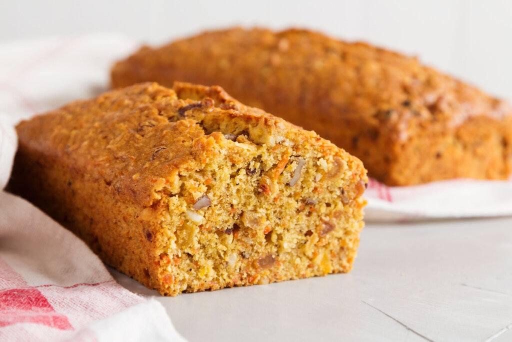 Bolo de cenoura e nozes cortada ao meio com bolo inteiro ao fundo em uma mesa branca