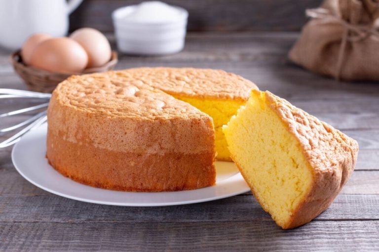 Bolo alto e macio feito com ovos, exibido em fatia sobre prato branco rústico com ingredientes ao fundo