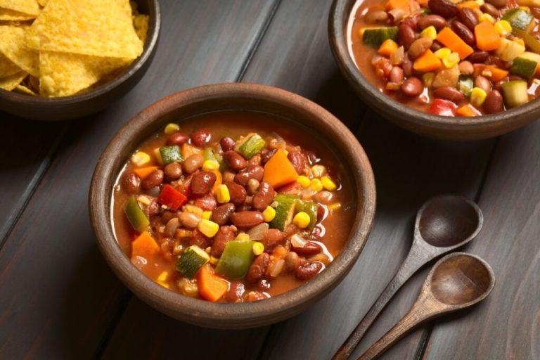Bowl de chili vegano com feijões, milho, cenoura, abobrinha e pimentões, servido em tigela de madeira ao lado de tortilhas e colheres rústicas