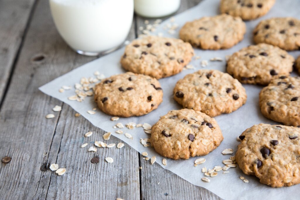 Cookies de aveia com gotas de chocolate dispostos sobre papel-manteiga, ao lado de copos de leite, com grãos de aveia espalhados