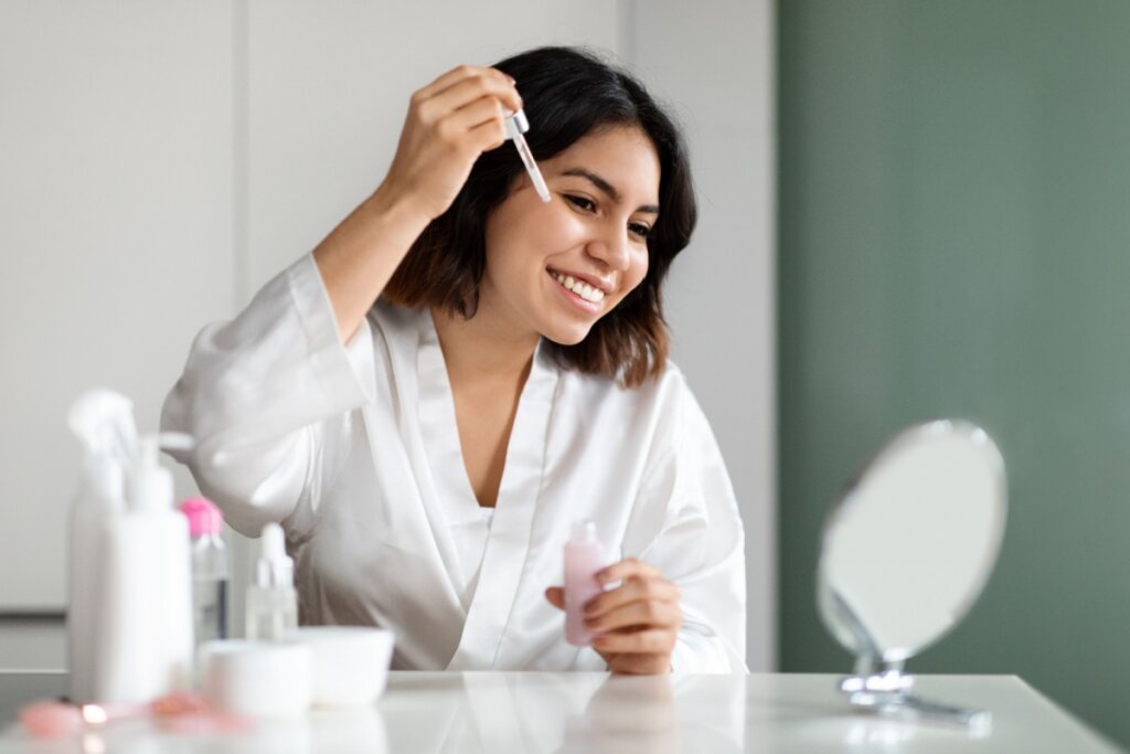 Mulher sorrindo, se olhando em um espelho e passando sérum no rosto