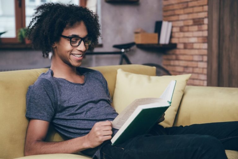 Homem de óculos e blusa cinza sentado em um sofá amarelo lendo livro 