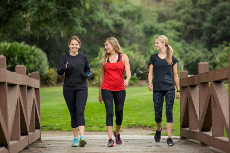 Três mulheres caminhando em ponte com árvores de parque ao fundo