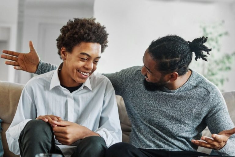 Jovem com o cabelo curto cacheado, usando camisa clara de botões conversando com o pai que está com o cabelo trançado e preso usando uma blusa de frio cinza sentados no sofá