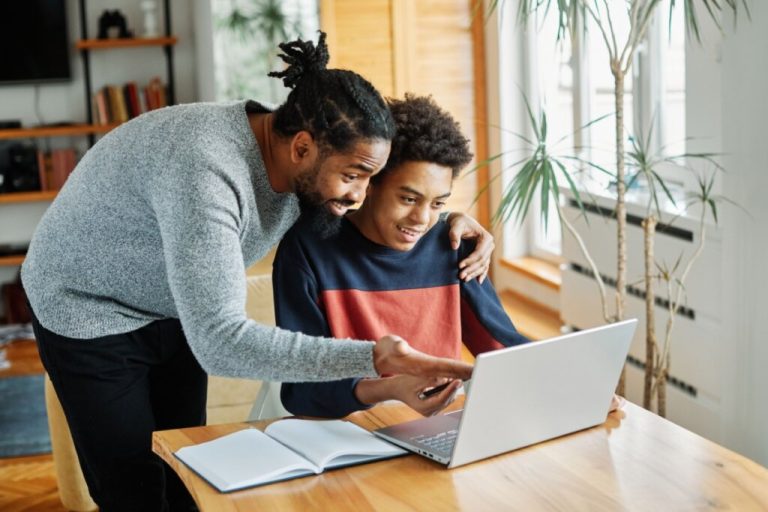 Pai ajudando filho a estudar, o filho tem o cabelo cacheado curto e está usando uma camisa azul e laranja, sentado em frente ao computador estudando e o pai está ao lado indicando algo no computador, ele tem o cabelo curto trançado e está usando uma camisa cinza e calça preta