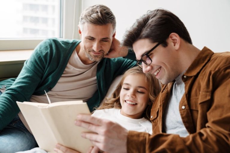 Pais lendo livro com filha sentados no sofa, um deles tem o cabelo curto e barba grisalha e está usando camisa bege e casaco verde, o outro tem o cabelo curto, está usando óculos preto, camisa cinza e camisa de botões marrom com um livro na mão, e no meio está a menina com o cabelo liso e claro, usando camiseta branca 