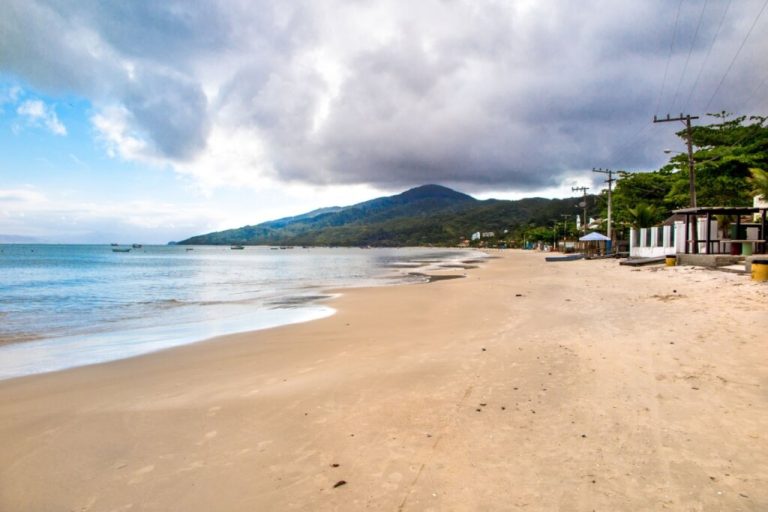 Foto da Praia de Zimbros, com extensa faixa de areia e águas cristalinas, com casas na beira da areia