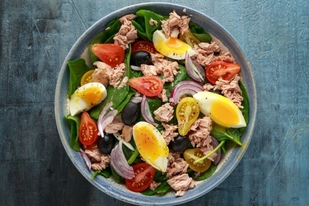 Salada de espinafre com atum, ovo, tomate, azeitonas pretas em cima de mesa de madeira azul
