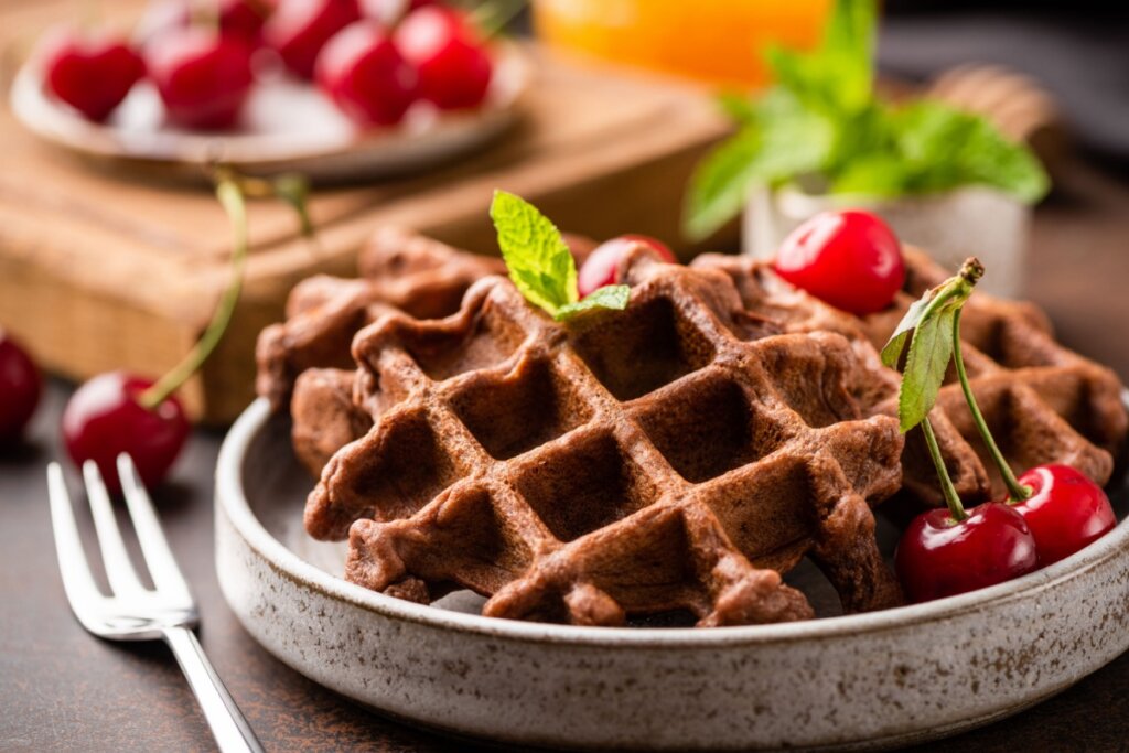 Waffle de chocolate em prato raso de cerâmica em cima de mesa de madeira, com garfo ao lado e cerejas